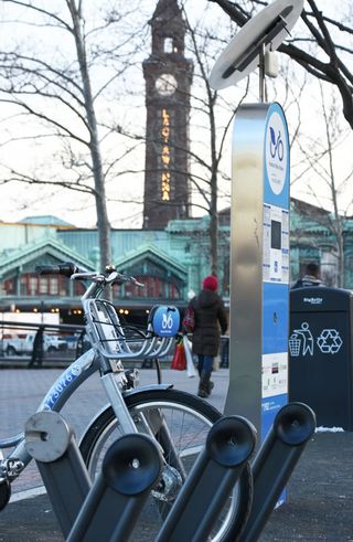 A silvery bicycle connected to a metal rack with a cable through the front wheel. In the foreground there are several empty docking slots, posts with circular connectors at the top. In the background is the waterfron walkway and Hoboken Terminal. A rental kiosk with a screen and solar panel is next to the bike.
